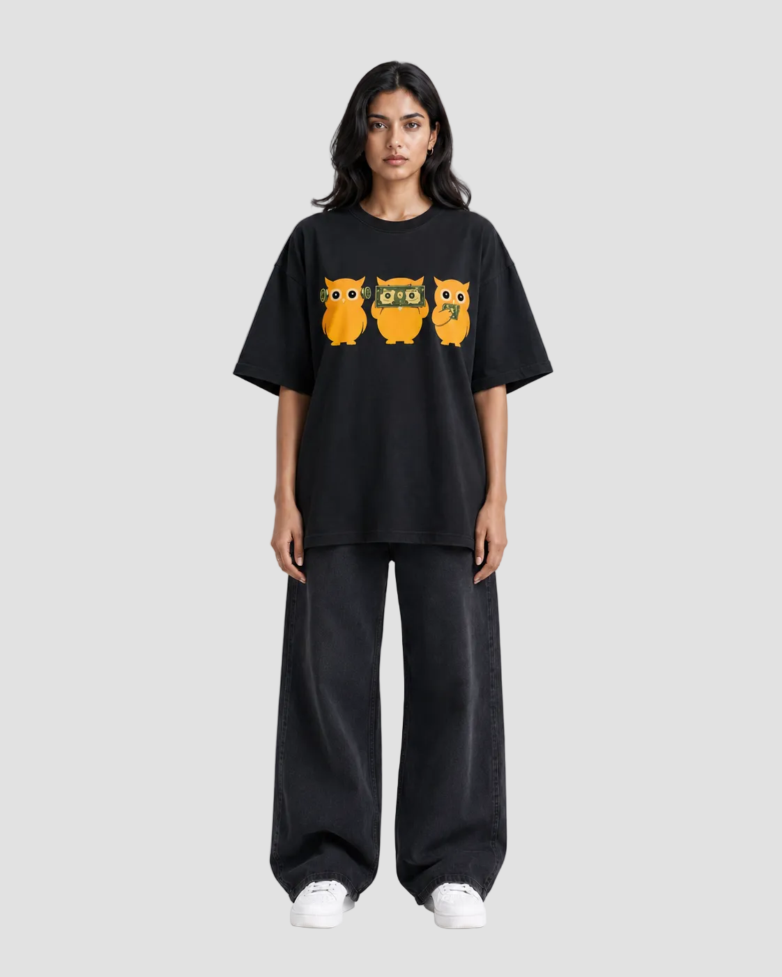 A model stands against a light gray studio background wearing a black oversized T-shirt with a graphic of three orange owls across the chest, paired with loose black pants and white sneakers.