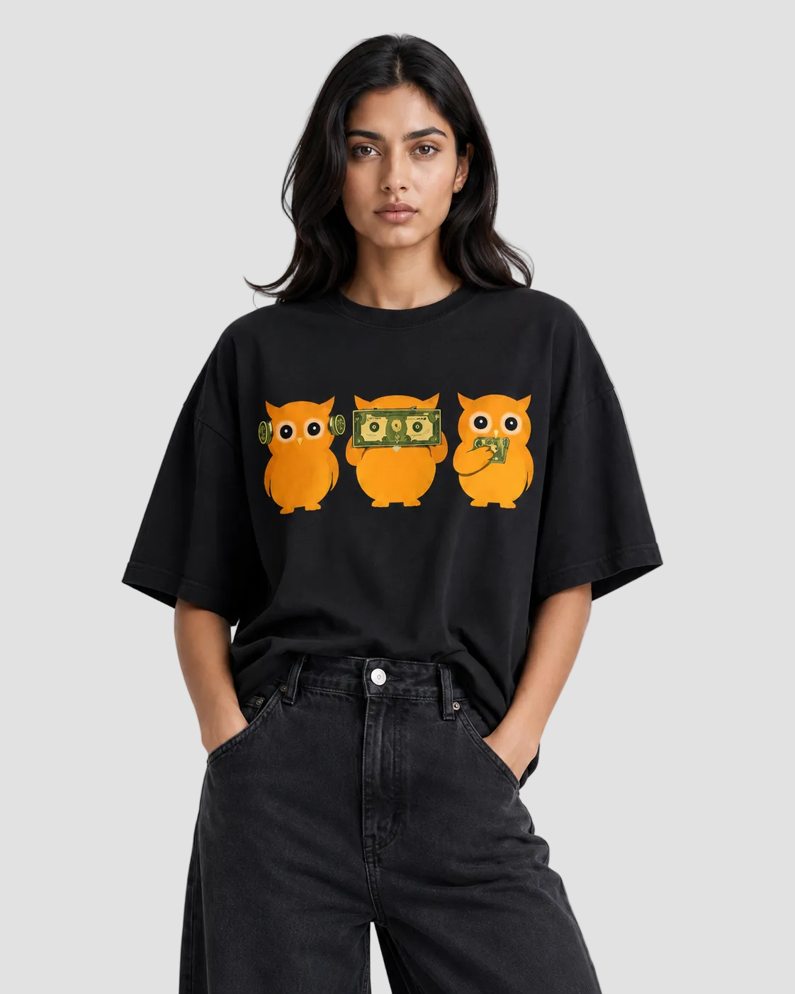 A woman modeling a black oversized t-shirt with a graphic of three orange owls wearing sunglasses, money bags, and a peace sign, paired with black jeans against a light gray background.