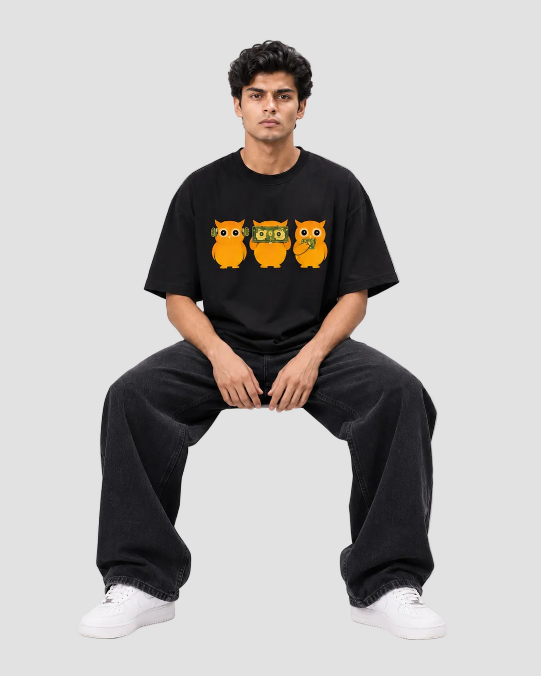 A man wearing a black t-shirt with a graphic of three orange owls, paired with black sweatpants and white sneakers, sitting on a white block against a light gray background.