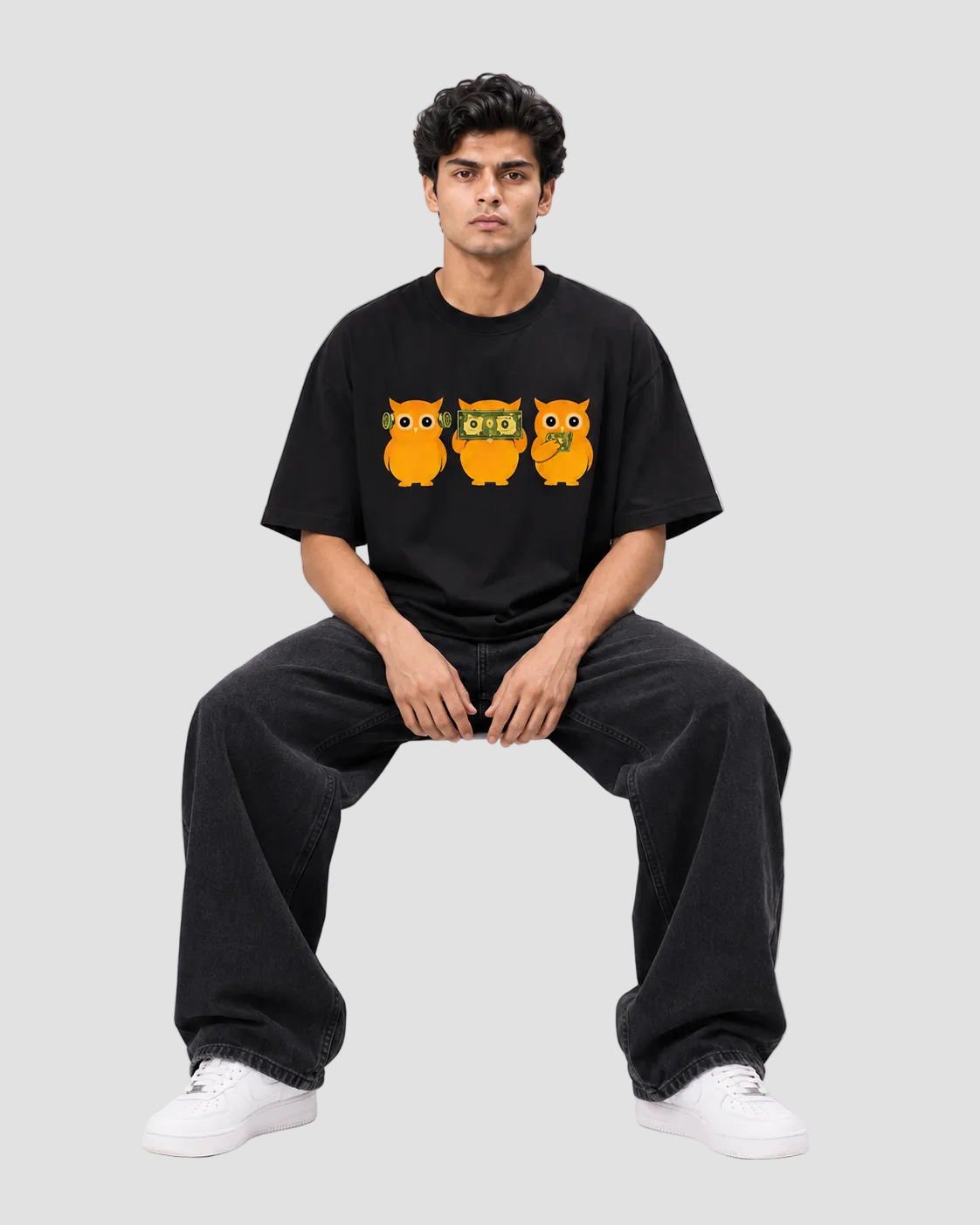 A man wearing a black t-shirt with a graphic of three orange owls, paired with black sweatpants and white sneakers, sitting on a white block against a light gray background.