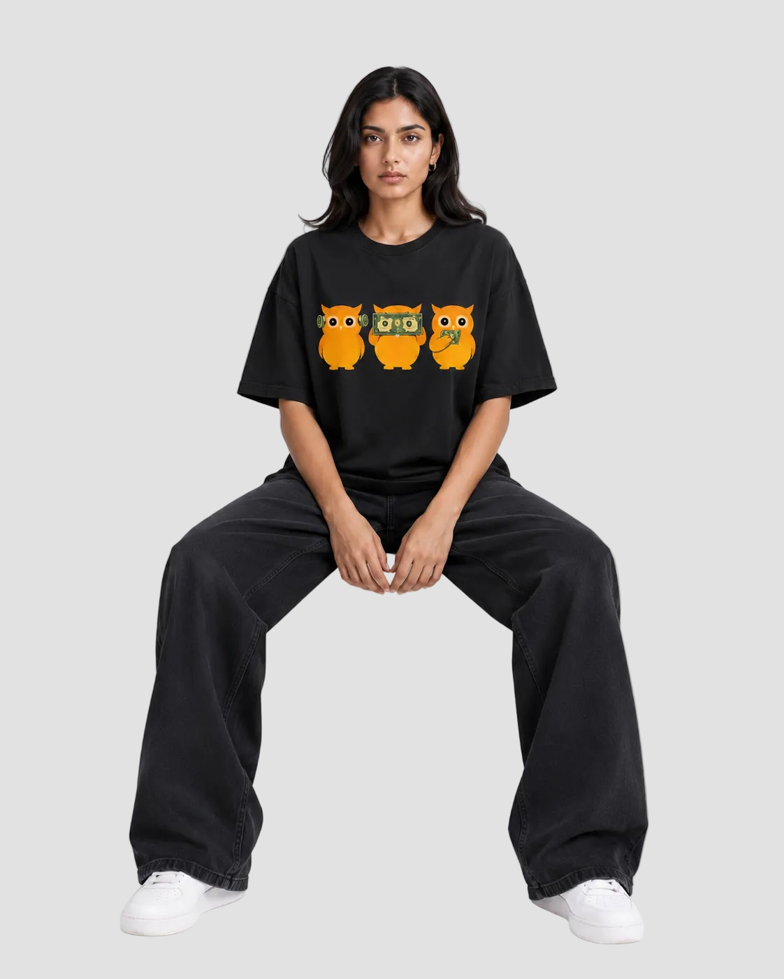 A woman sits on a white block wearing a black oversized T-shirt with three cartoon owls across the chest, black wide-leg pants, and white sneakers against a plain light gray studio background.