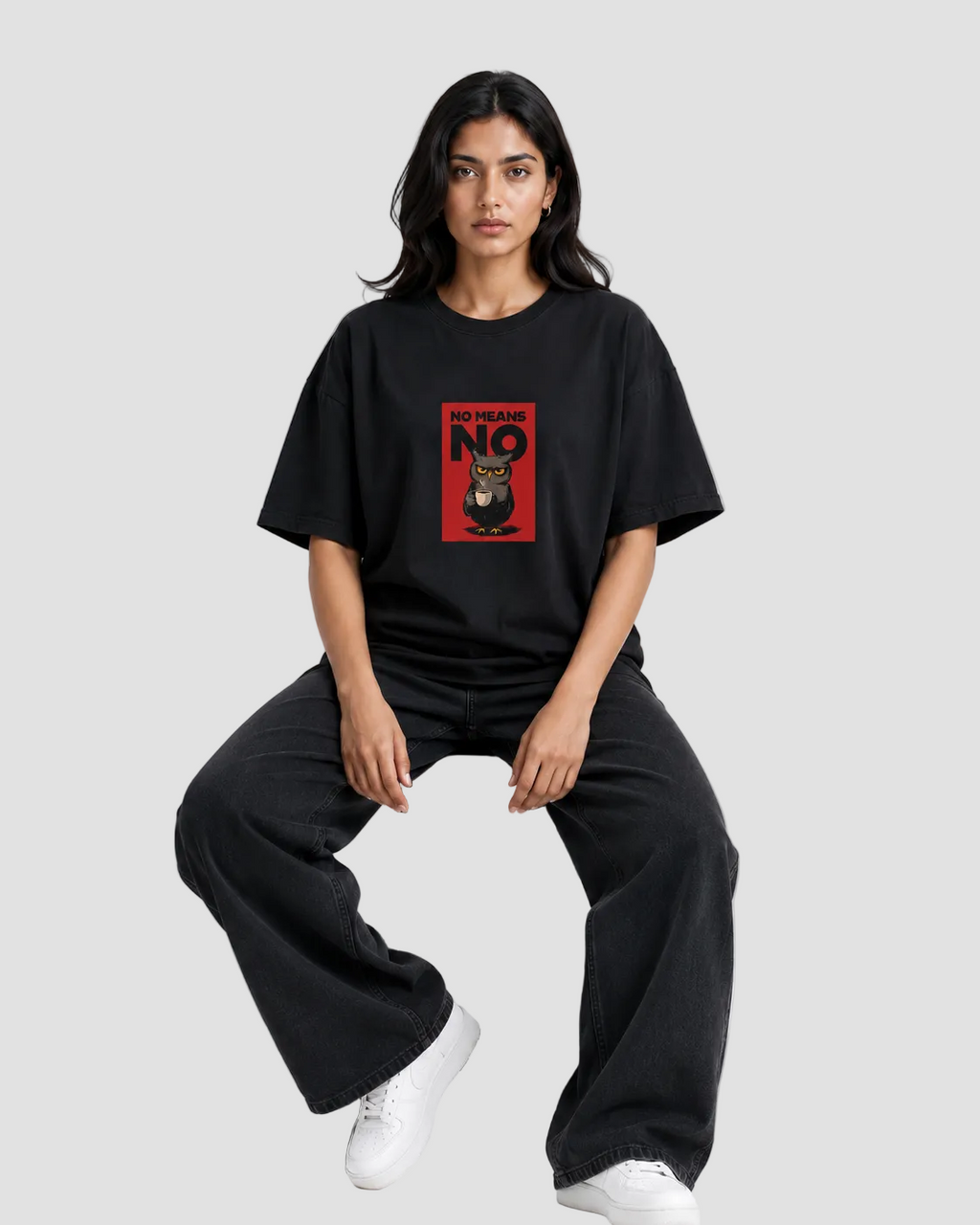 A woman sits on a black stool wearing an oversized black t-shirt with a red graphic print on the chest, paired with black wide-leg sweatpants and white sneakers, against a light gray studio background.
