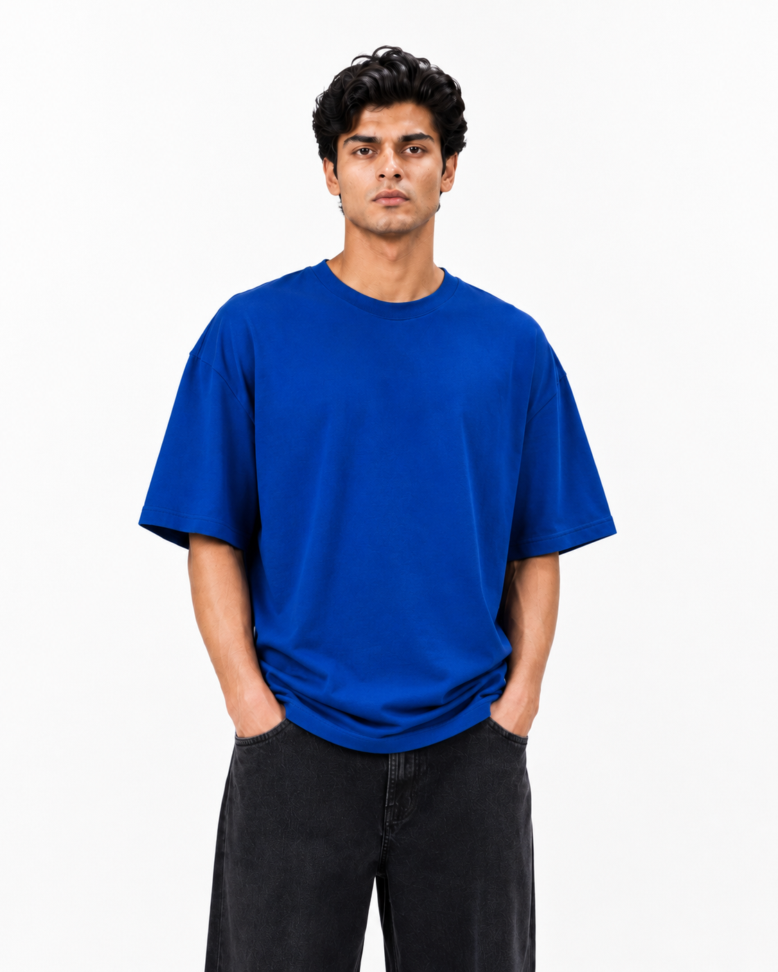 A man wearing a plain royal blue short-sleeve crewneck t-shirt and dark gray jeans, standing against a light gray studio background.