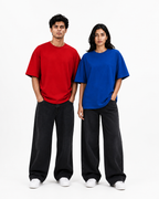 Two models standing side-by-side wearing oversized short-sleeve t-shirts in red and blue, paired with black wide-leg jeans and white sneakers, against a plain white studio background.