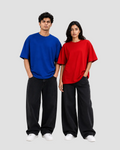 A man and a woman modeling oversized short-sleeve t-shirts in vibrant royal blue and bright red, paired with loose black pants and white sneakers, against a plain white studio background.