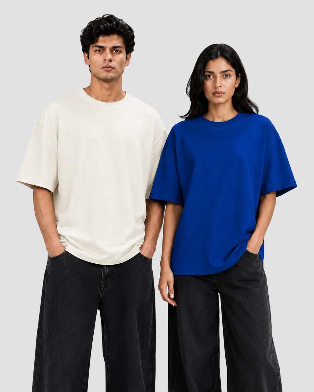 A man and a woman modeling oversized short-sleeve crewneck t-shirts. The man wears an off-white/cream t-shirt and dark charcoal pants, while the woman wears a vibrant royal blue t-shirt and dark charcoal pants.