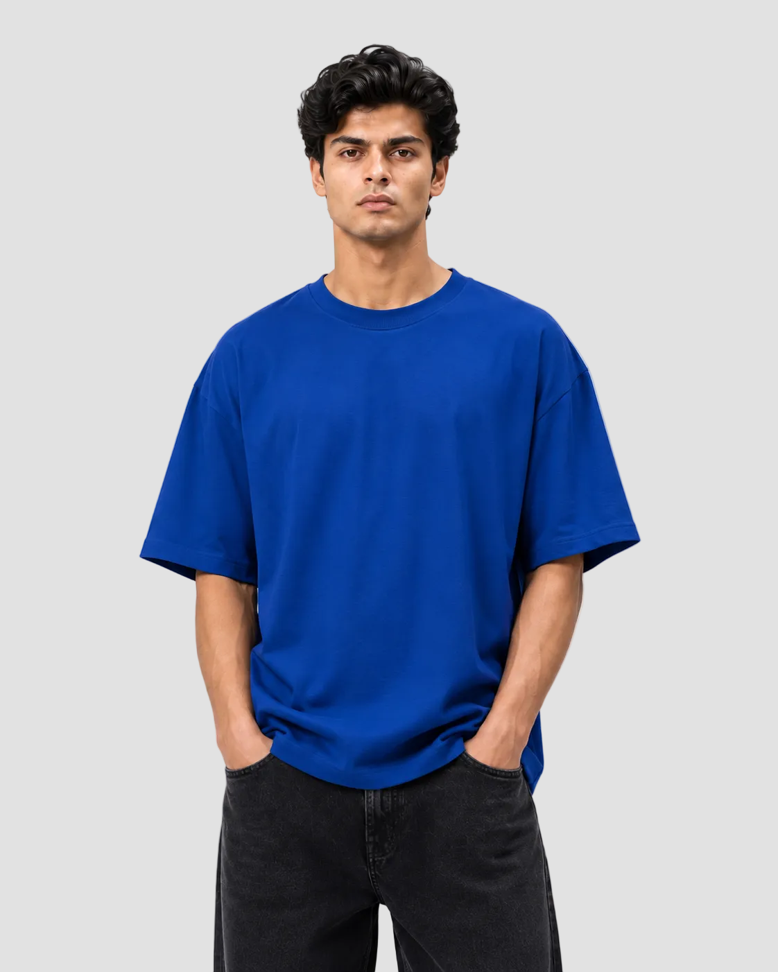 A man wearing a plain royal blue short-sleeve crewneck t-shirt and black jeans, standing against a light gray studio background.