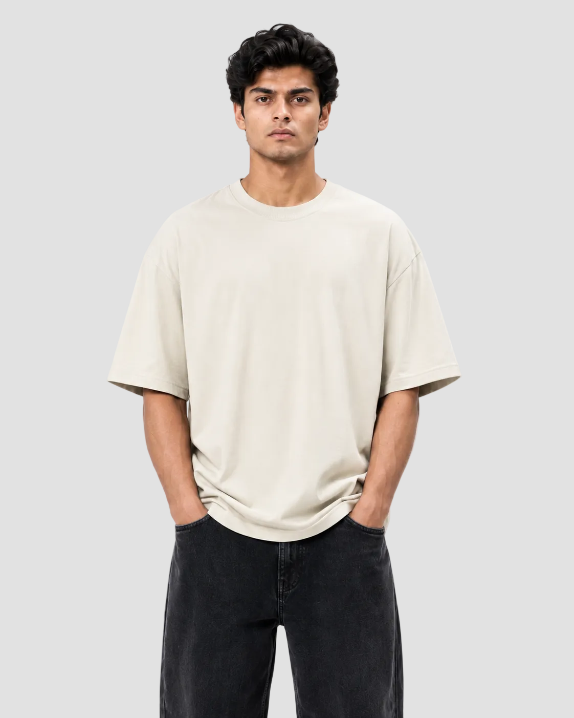 A man wearing a light beige crew-neck t-shirt and dark charcoal pants, standing against a plain white studio background.