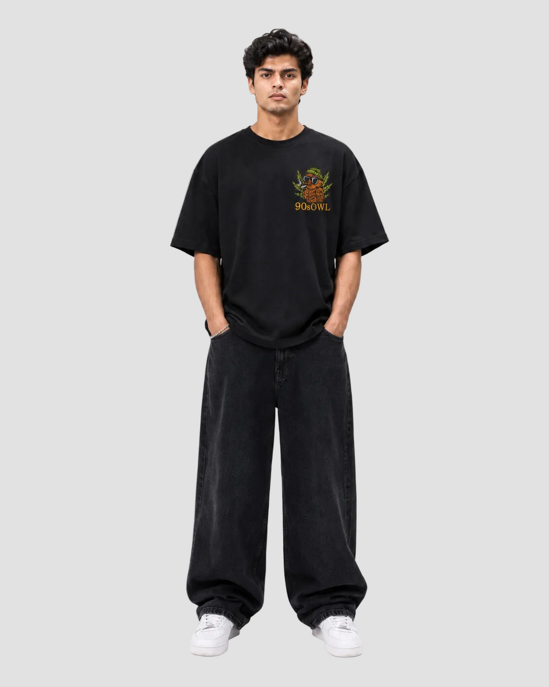 A man modeling a black crewneck t-shirt with a colorful graphic on the left chest that reads 'BOYBLOOD' and '1984', paired with black wide-leg sweatpants and white sneakers, against a plain light gray background.