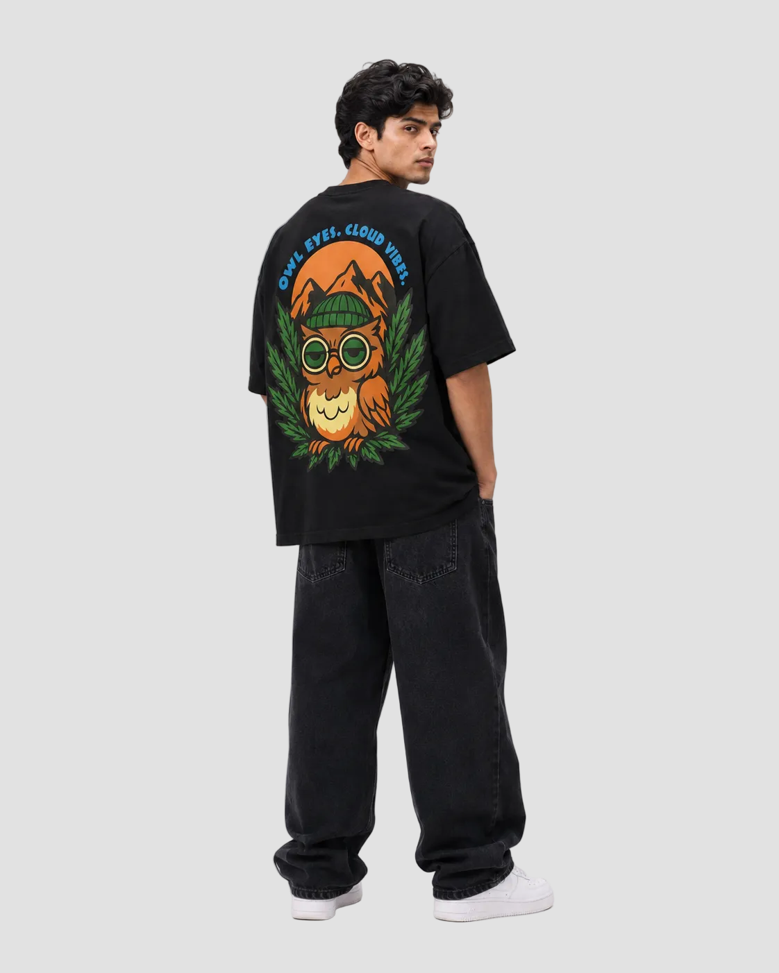 A man wearing a black t-shirt with a large colorful owl graphic on the back, paired with black jeans and white sneakers, photographed from behind against a light gray studio background.