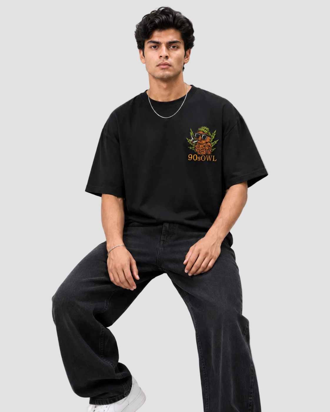 A man sitting on a black stool wearing a black oversized t-shirt with a colorful 90GOWL chest graphic, paired with black wide-leg jeans and white sneakers, against a light gray background.