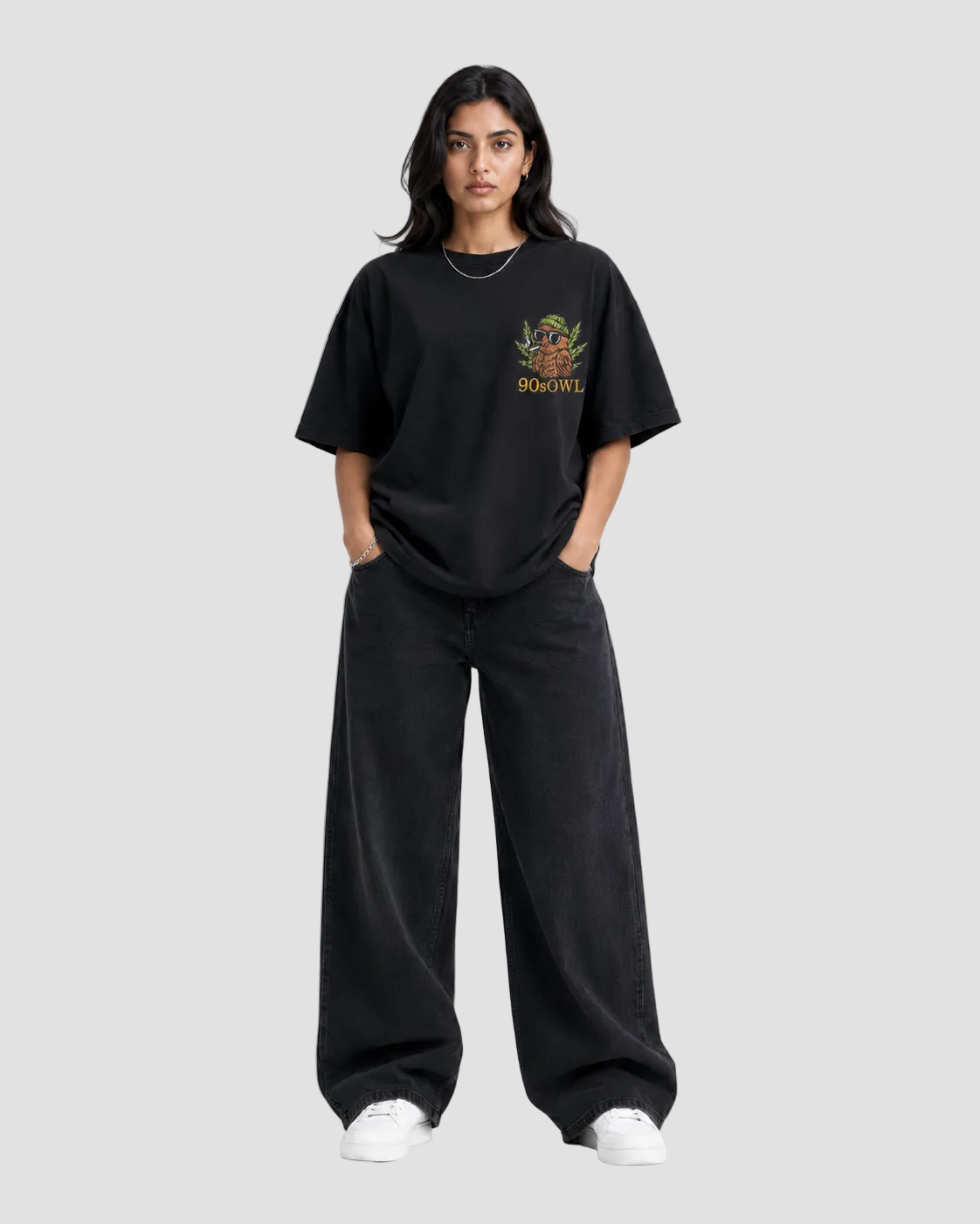 A woman modeling a black oversized t-shirt with a small multicolor graphic on the left chest and matching black wide-leg pants, standing against a light gray studio background.
