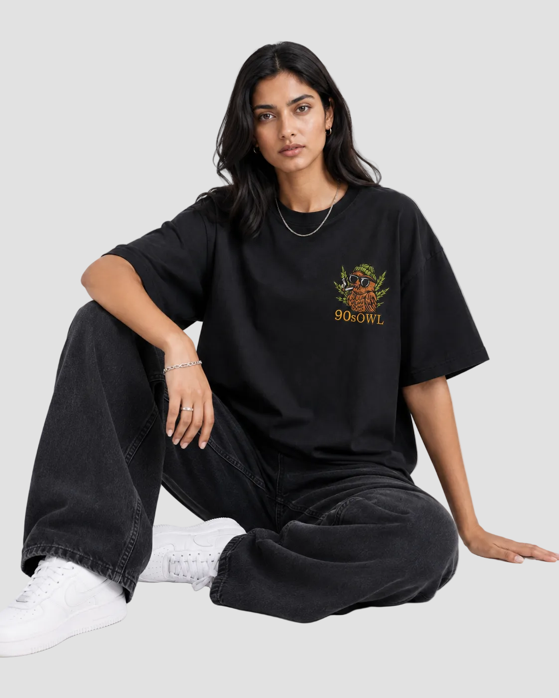 A woman sits cross-legged on a light gray studio floor wearing an oversized black graphic T-shirt with a colorful chest print reading 'SOKOWY' and dark wide-leg jeans with white sneakers.