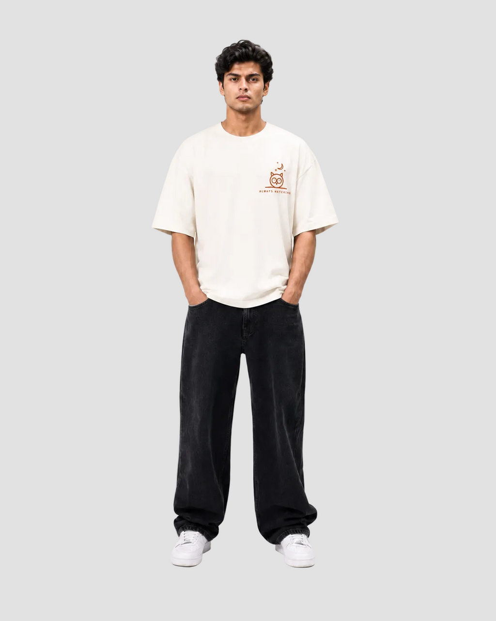 A man modeling a cream-colored short-sleeve t-shirt with a small orange graphic on the left chest, paired with black wide-leg pants and white sneakers, against a plain white studio background.