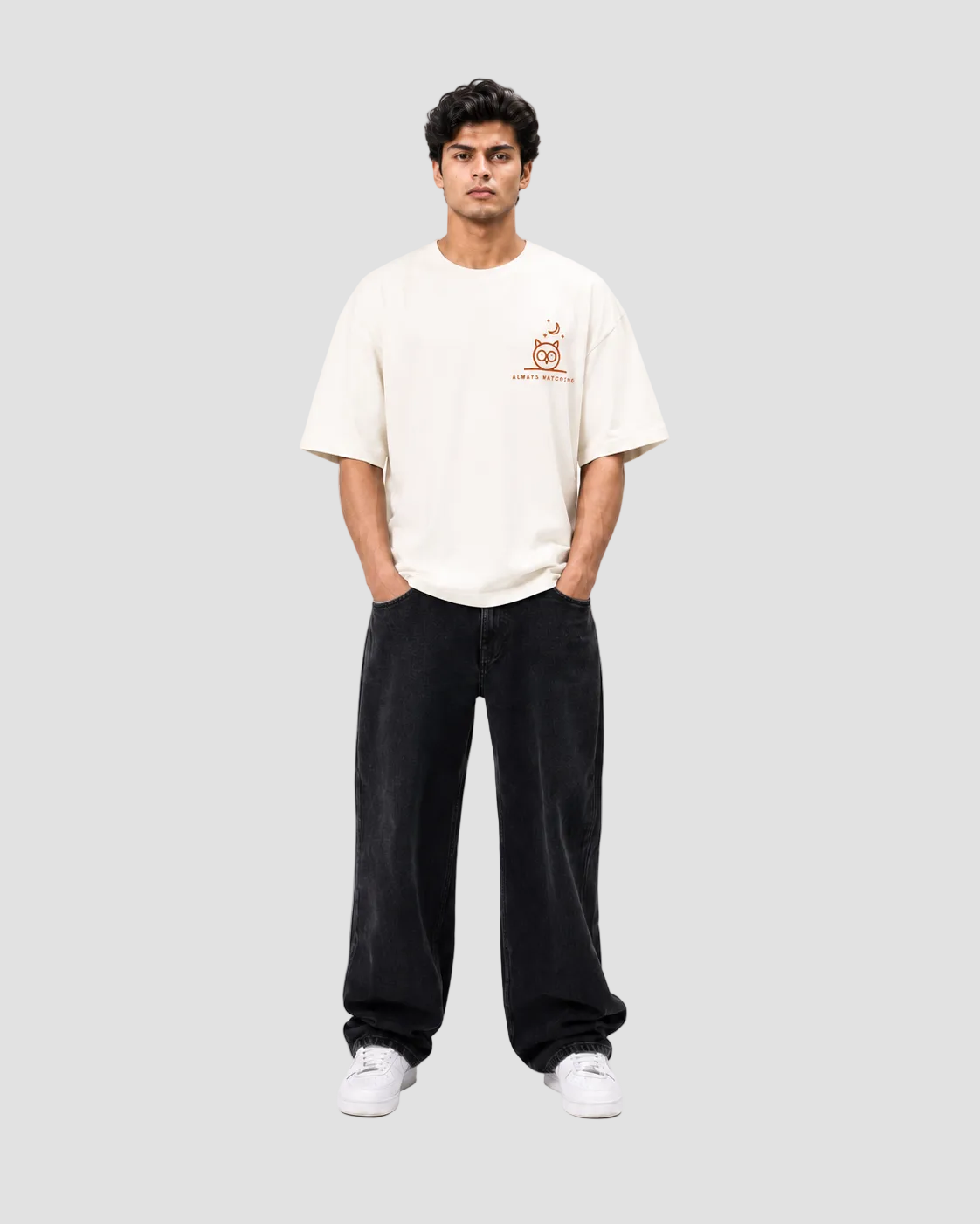 A man modeling a cream-colored short-sleeve t-shirt with a small orange graphic on the left chest, paired with black wide-leg pants and white sneakers, against a plain white studio background.