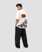 A man wearing a cream-colored oversized t-shirt with a graphic print of black and white owls and a brown speech bubble on the back, paired with dark charcoal jeans and white sneakers, photographed against a seamless white studio background.