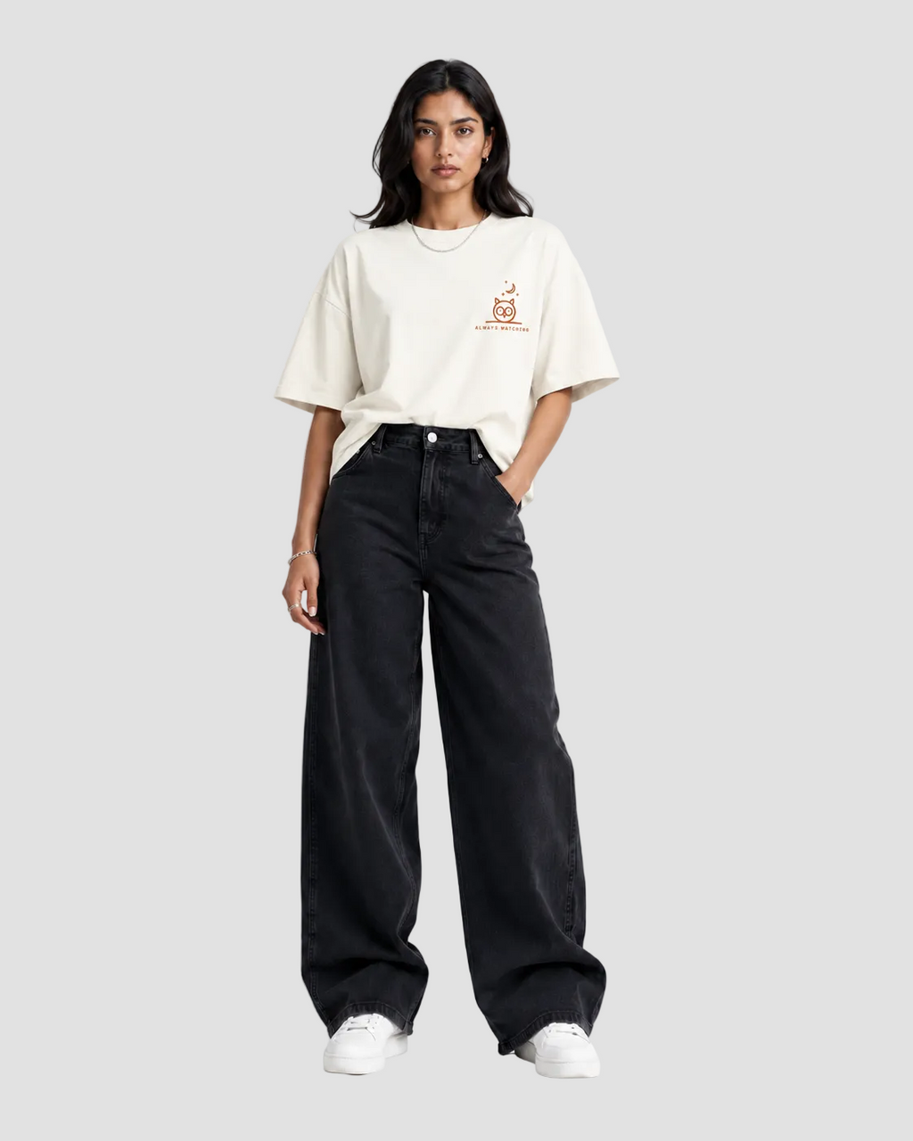 A woman stands against a light gray studio background wearing a cream-colored oversized t-shirt with a small orange graphic on the left chest, paired with black wide-leg jeans and white sneakers.