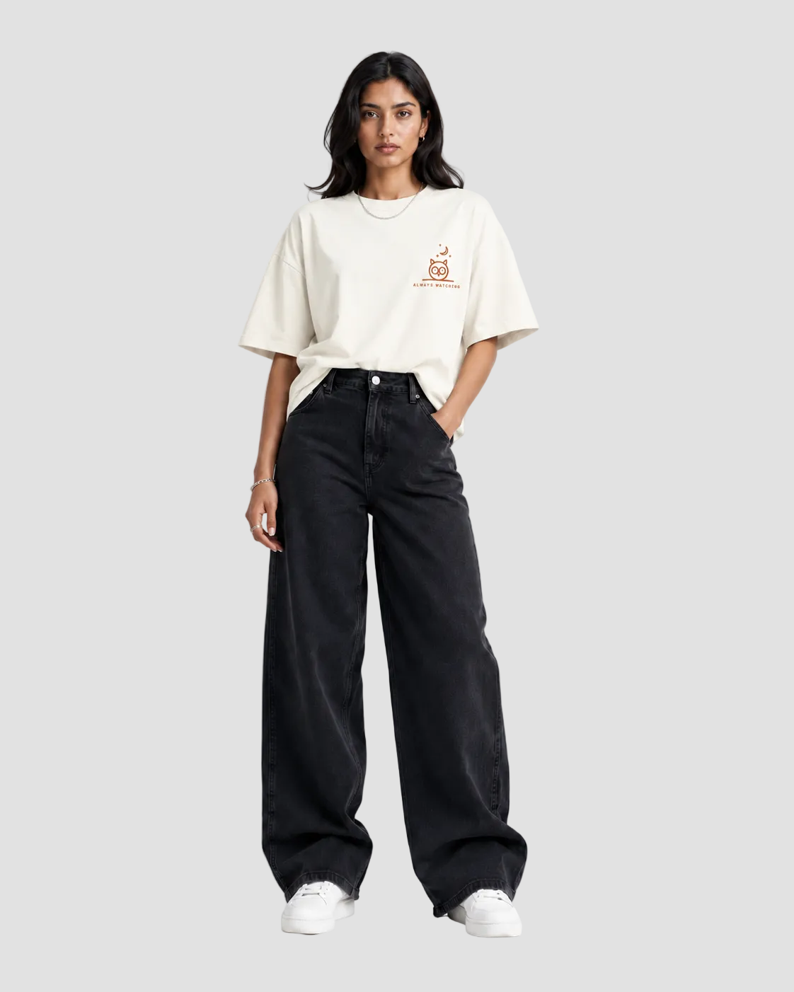 A woman stands against a light gray studio background wearing a cream-colored oversized t-shirt with a small orange graphic on the left chest, paired with black wide-leg jeans and white sneakers.