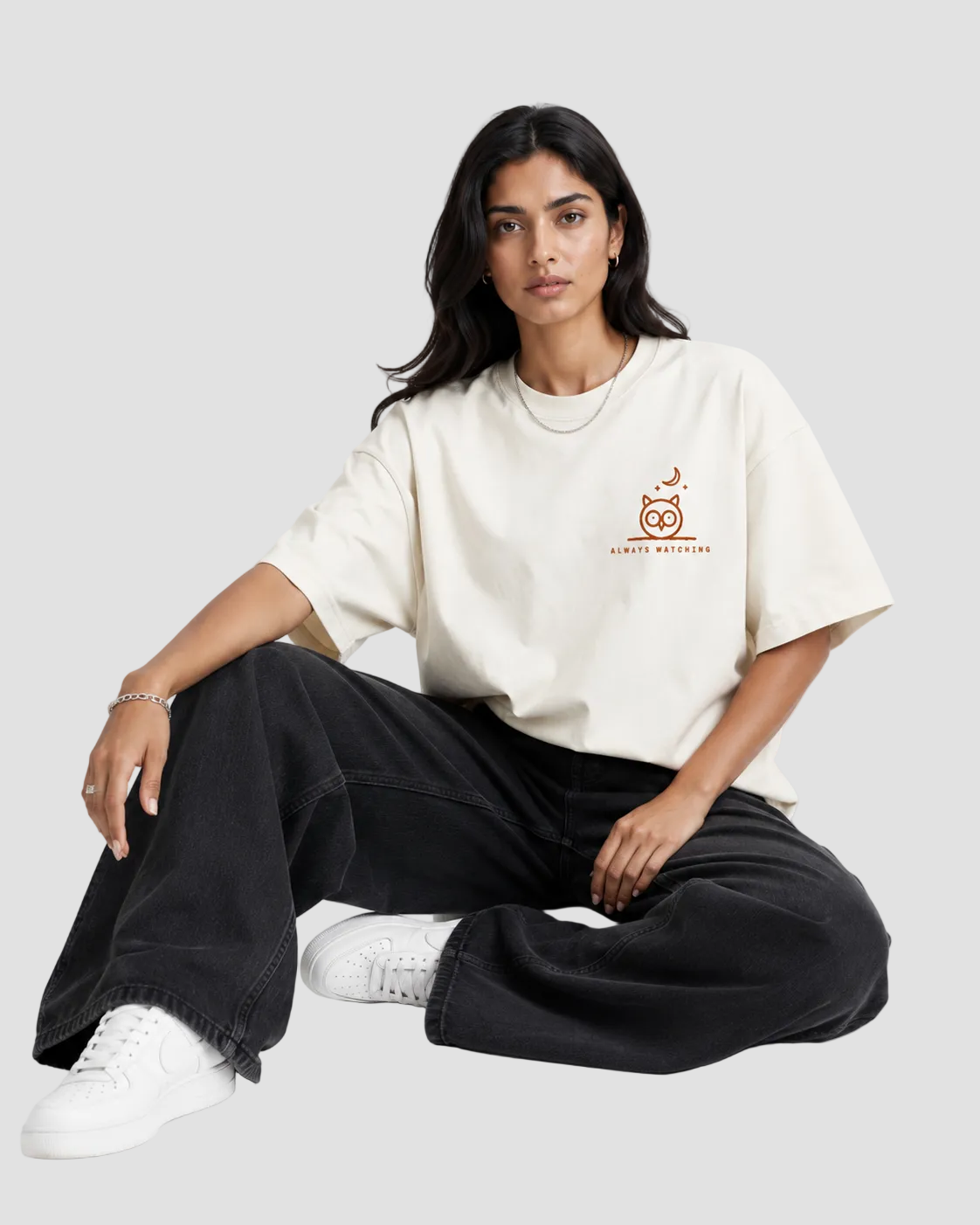 A woman sits cross-legged on a light gray studio floor wearing an oversized off-white t-shirt with a small orange graphic on the left chest, black wide-leg jeans, and white sneakers.