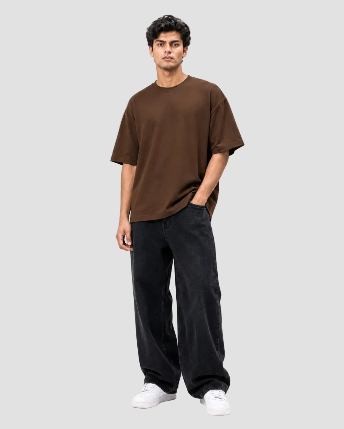A man modeling an oversized brown short-sleeve t-shirt, paired with loose black jeans and white sneakers, standing against a plain white studio background.
