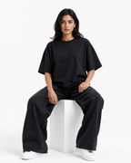 A woman sits on a white cube in a studio setting, wearing an oversized black short-sleeve t-shirt, matching black wide-leg pants, and white sneakers.
