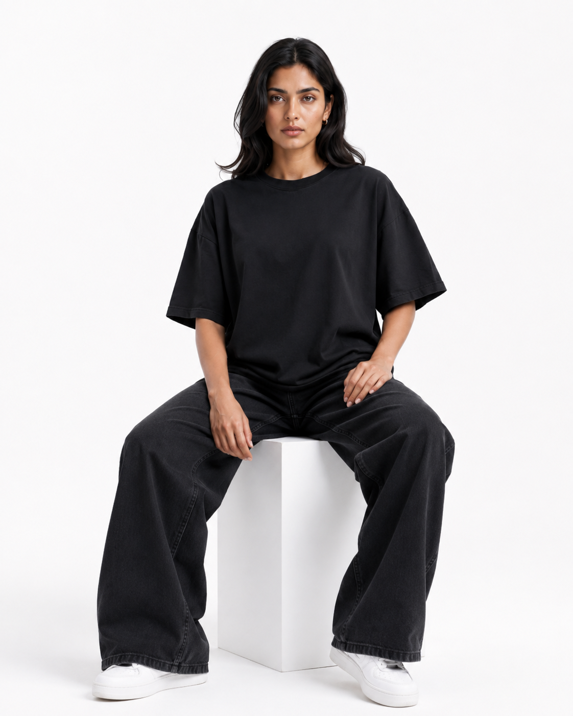 A woman sits on a white cube in a studio setting, wearing an oversized black short-sleeve t-shirt, matching black wide-leg pants, and white sneakers.