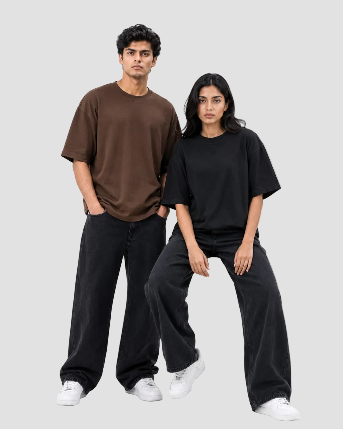 A man and a woman modeling oversized short-sleeve t-shirts and wide-leg jeans against a plain white studio background. The man wears an oversized brown t-shirt and dark jeans, while the woman wears an oversized black t-shirt and dark jeans, both paired with white sneakers.