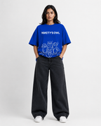 A model stands against a light gray studio background wearing a bright blue graphic T-shirt with the text 'NINETY'S OWL' and white cartoon-style line art, paired with loose dark charcoal pants and white sneakers.