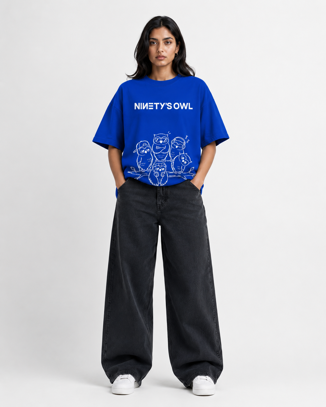 A model stands against a light gray studio background wearing a bright blue graphic T-shirt with the text 'NINETY'S OWL' and white cartoon-style line art, paired with loose dark charcoal pants and white sneakers.