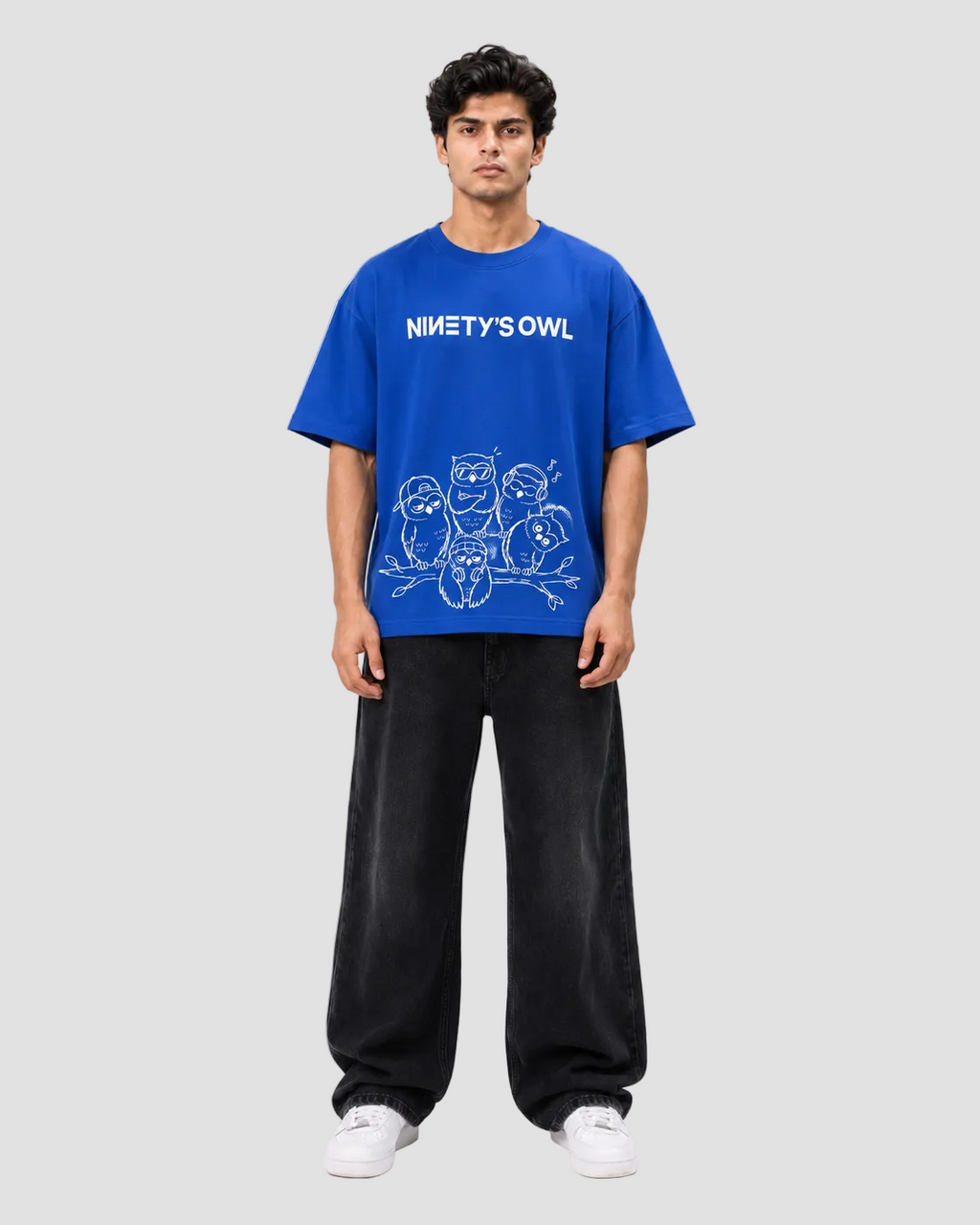 A man modeling a vibrant blue t-shirt with a white graphic print that reads 'NINETY'S OWL' above cartoon owl illustrations. He is wearing loose black sweatpants and white sneakers against a plain light gray studio background.