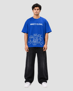 A man modeling a vibrant blue t-shirt with a white graphic print that reads 'NINETY'S OWL' above cartoon owl illustrations. He is wearing loose black sweatpants and white sneakers against a plain light gray studio background.