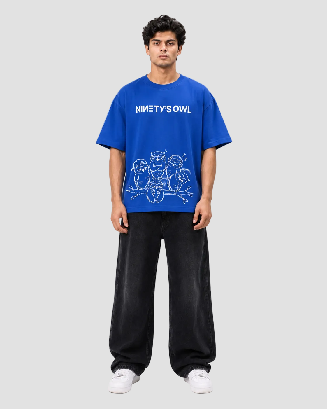 A man modeling a vibrant blue t-shirt with a white graphic print that reads 'NINETY'S OWL' above cartoon owl illustrations. He is wearing loose black sweatpants and white sneakers against a plain light gray studio background.