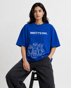 A woman wearing an oversized royal blue t-shirt with 'NINETY'S OWL' and an owl graphic printed in white, paired with dark gray wide-leg jeans, sitting on a black stool against a light gray background.