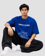 A man sitting cross-legged wearing a royal blue t-shirt with 'NINETY'S OWL' printed in white across the chest, paired with black jeans and white sneakers, against a light gray studio background.