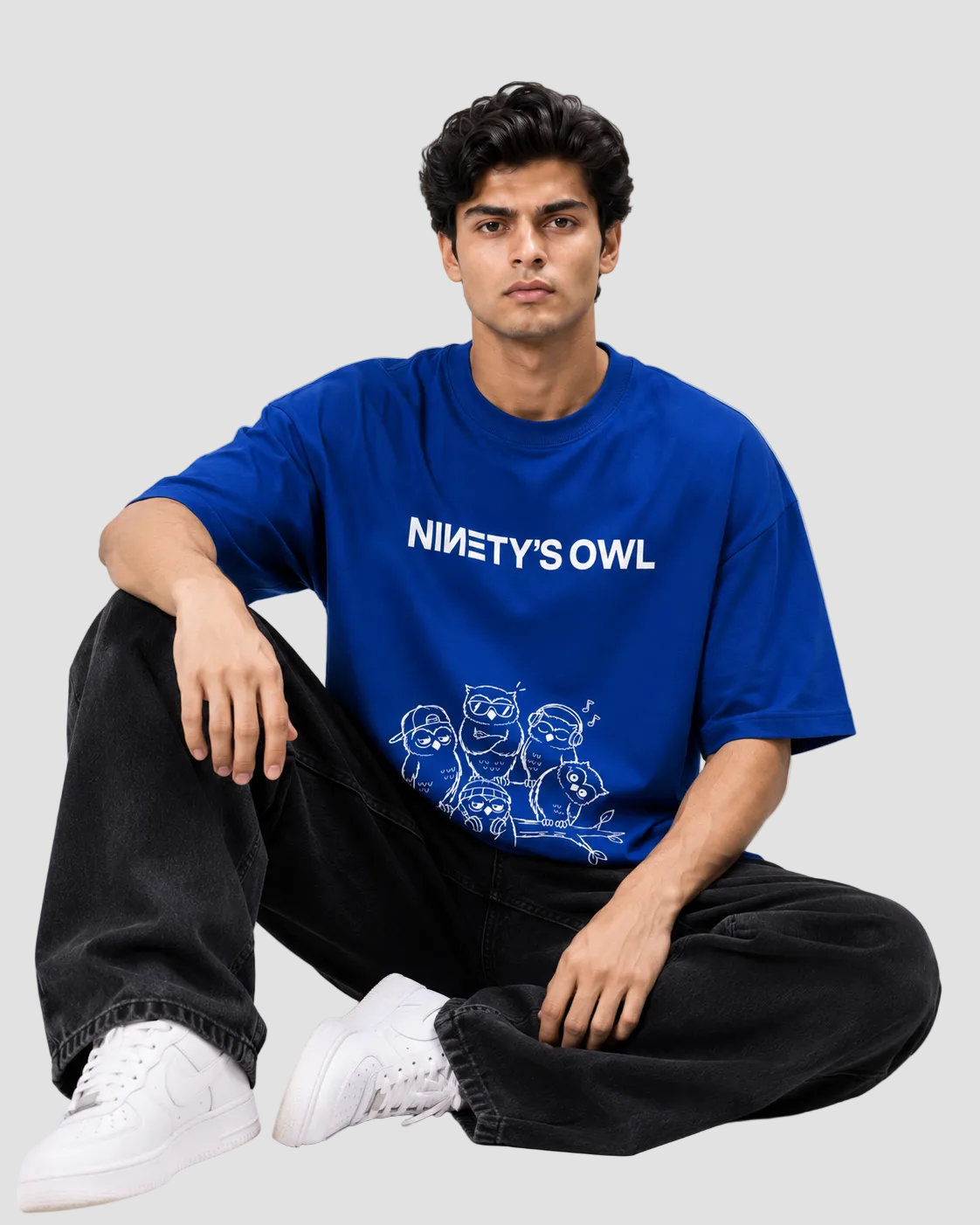 A man sitting cross-legged wearing a royal blue t-shirt with 'NINETY'S OWL' printed in white across the chest, paired with black jeans and white sneakers, against a light gray studio background.