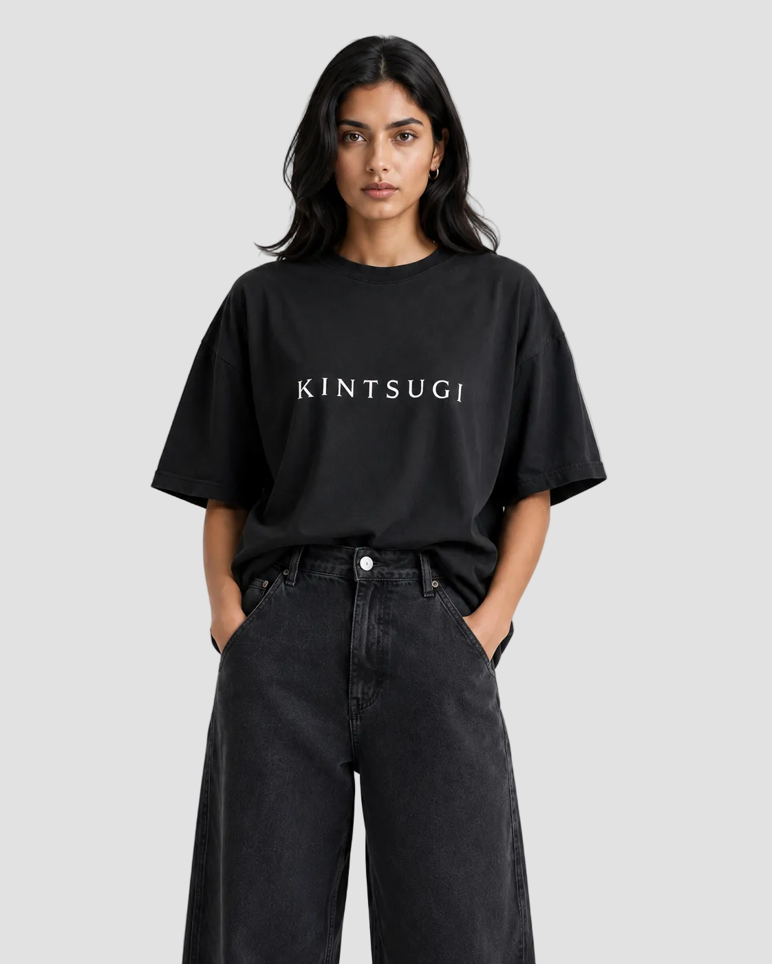 A woman modeling an oversized black t-shirt with 'KINTSUGI' printed in white across the chest, paired with dark wide-leg jeans against a light gray studio background.