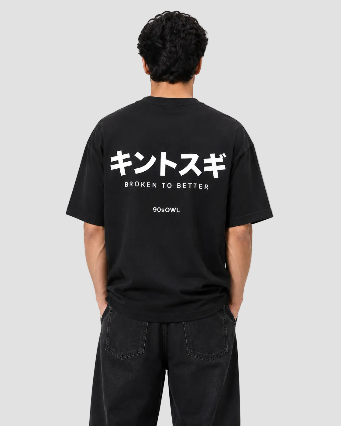 A man viewed from behind wearing a black t-shirt with white Japanese text and 'BROKEN TO BETTER' printed on the back, along with the brand '901OWL'.