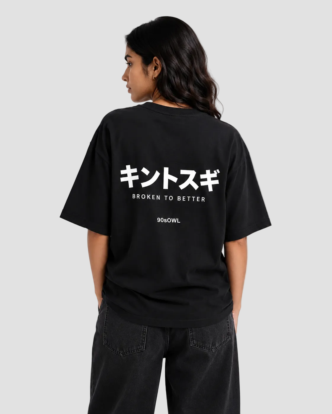 A woman shown from the back wearing a black oversized t-shirt with white Japanese text and 'BROKEN TO BETTER' printed on the back, paired with black jeans against a light gray studio background.