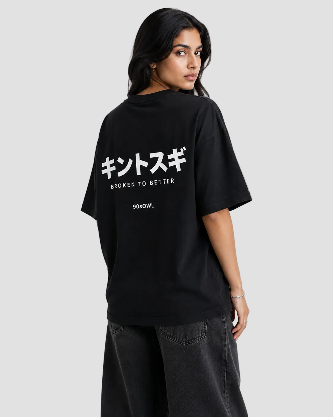 A man sitting on a stool wearing a black oversized t-shirt with 'KINTSUGI' printed in white across the chest, paired with dark gray jeans, against a light gray background.