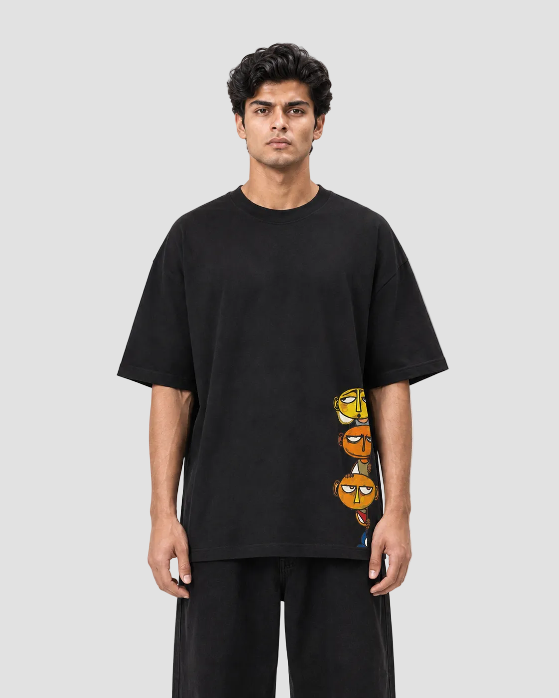A man wearing a black oversized short-sleeve t-shirt with a vertical graphic print of three cartoon orange characters on the lower right side, paired with black pants, against a plain white studio background.