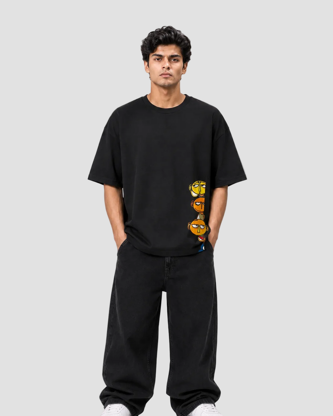A man wearing a black oversized t-shirt with a vertical graphic of three cartoon-style yellow and orange eyes, paired with black sweatpants, standing against a light gray studio background.