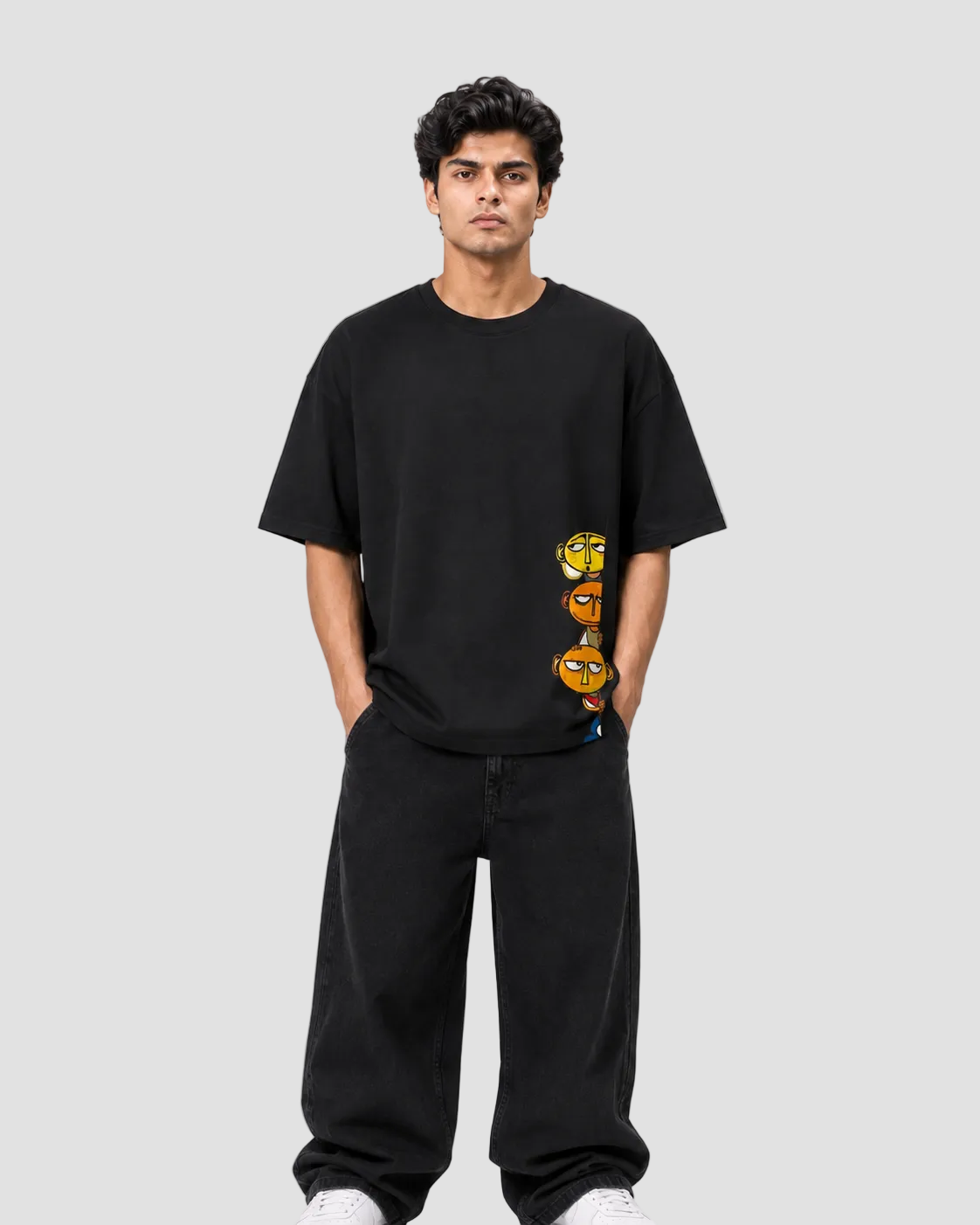 A man wearing a black oversized t-shirt with a vertical graphic of three cartoon-style yellow and orange eyes, paired with black sweatpants, standing against a light gray studio background.