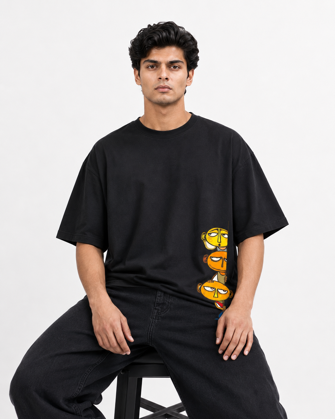 A man wearing a black oversized t-shirt with a vertical graphic of three cartoon faces in yellow, orange, and green, paired with black jeans, seated on a stool against a light gray background.