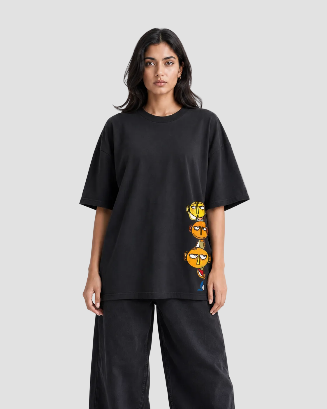 A woman modeling an oversized black short-sleeve t-shirt with a vertical graphic print of three cartoon characters on the lower right side, paired with black wide-leg pants against a plain white studio background.