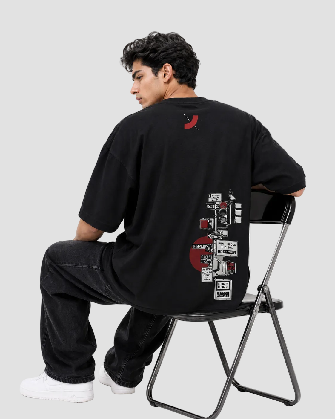 A man wearing an oversized black graphic t-shirt, black jeans, and white sneakers sits on a black folding chair, viewed from behind to show the back print of the t-shirt.