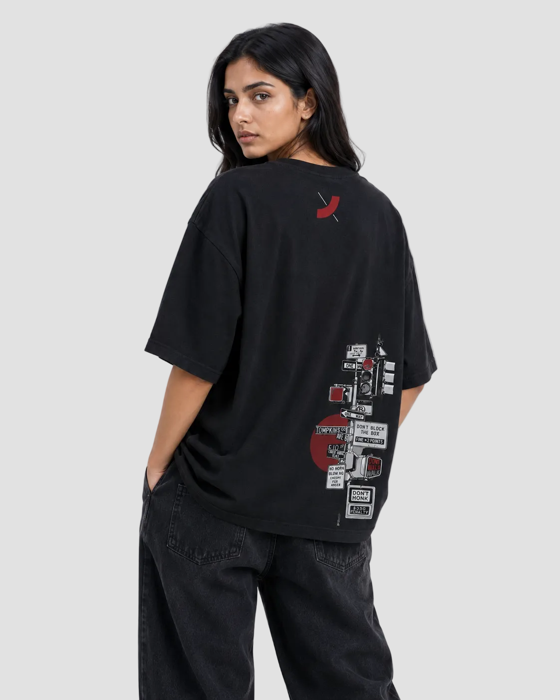 A woman wearing an oversized black graphic T-shirt, shown from the back against a plain light gray studio background. The shirt features a small red logo near the upper back and a large collage-style graphic print in red, white, and black along the lower back.