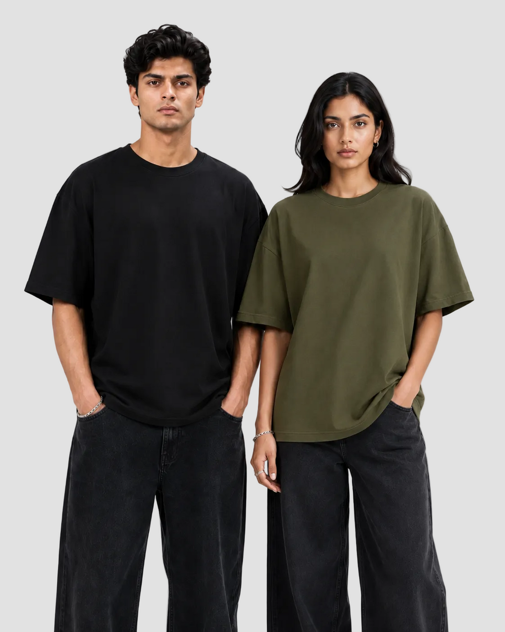 A man and a woman modeling oversized short-sleeve crewneck t-shirts against a plain light gray studio background. The man wears a black t-shirt and dark jeans, while the woman wears an olive green t-shirt and dark jeans.