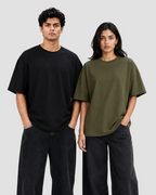 A man and a woman modeling oversized short-sleeve crewneck t-shirts against a plain light gray studio background. The man wears a black t-shirt and dark jeans, while the woman wears an olive green t-shirt and dark jeans.