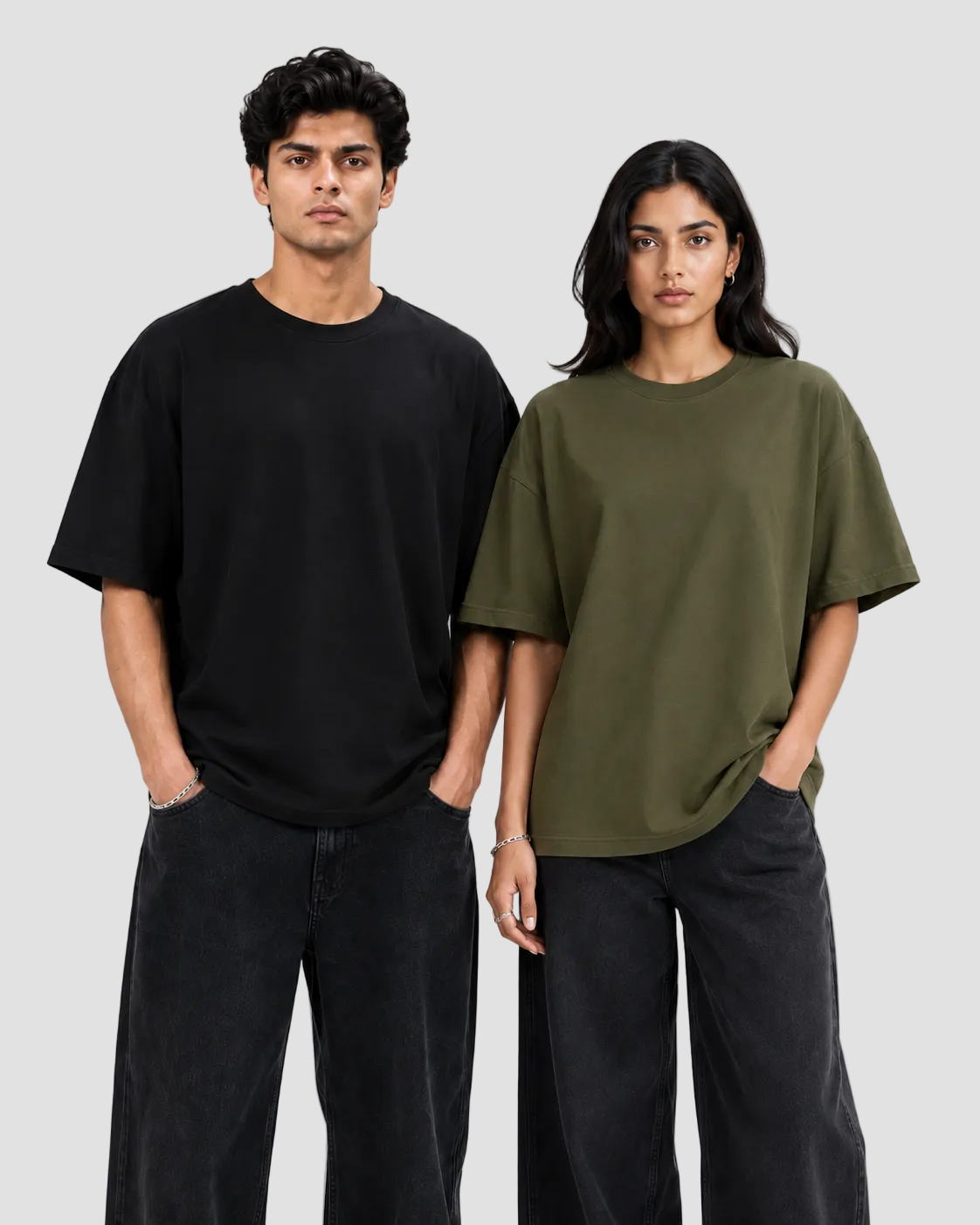 A man and a woman modeling oversized short-sleeve crewneck t-shirts against a plain light gray studio background. The man wears a black t-shirt and dark jeans, while the woman wears an olive green t-shirt and dark jeans.