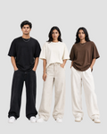 Three models standing side-by-side wearing oversized short-sleeve t-shirts in black, cream, and brown, paired with wide-leg trousers in black, beige, and off-white, against a plain white studio background.